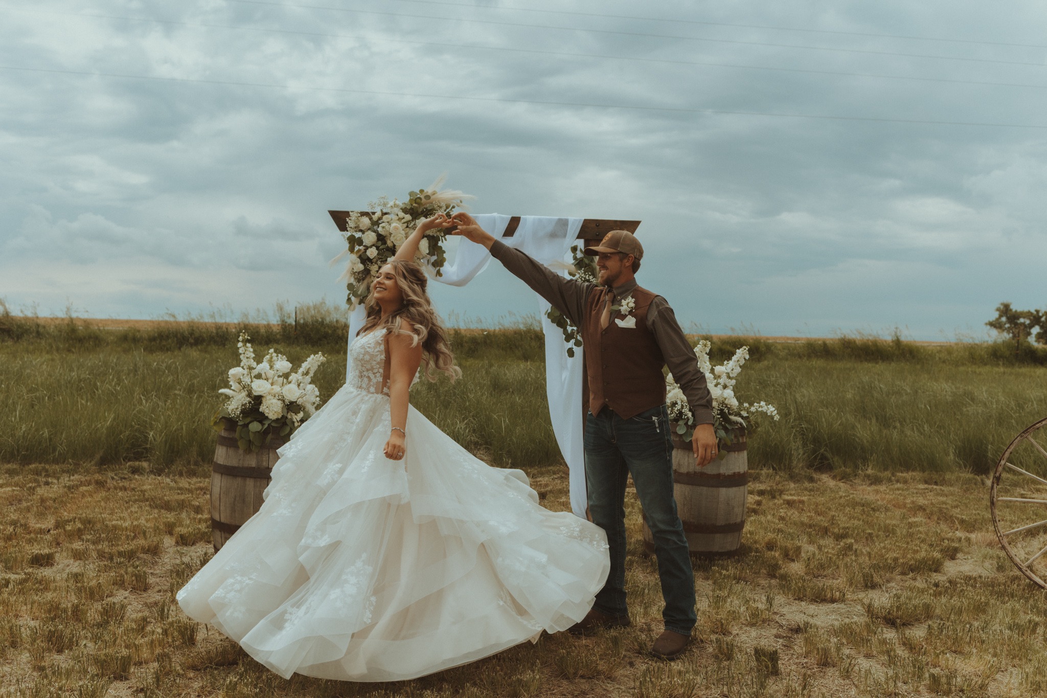 Derek twirling Jordan beneath their flower-covered wedding arch in the pasture